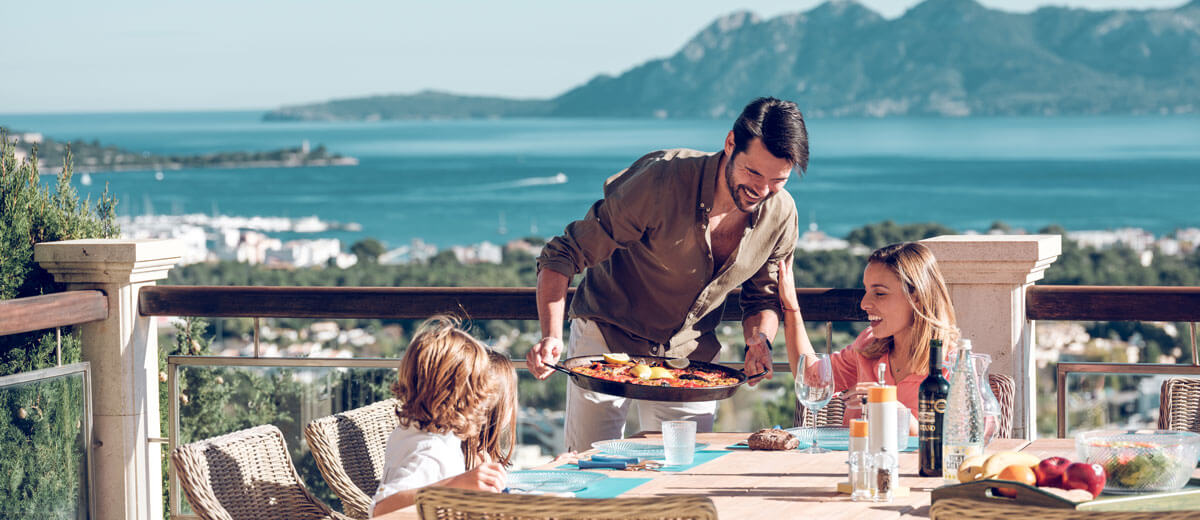 Familia disfrutando de una comida al aire libre en una terraza soleada con impresionantes vistas al mar y a la montaña: el padre sirve la comida mientras la madre y el niño sonríen en la mesa.